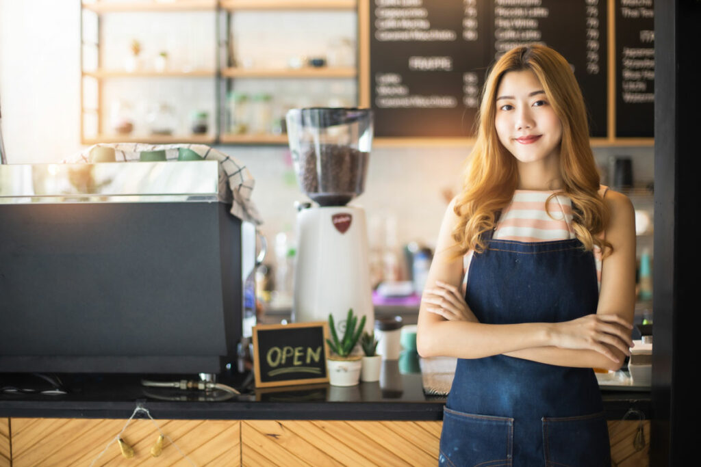 Pretty young asian waitress standing arms crossed in cafeteria.Coffee Business owner Concept. barista in apron smiling at camera in coffee shop counter