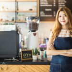 Pretty young asian waitress standing arms crossed in cafeteria.Coffee Business owner Concept. barista in apron smiling at camera in coffee shop counter